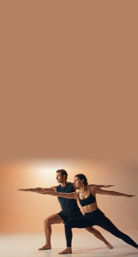 A man and a woman in athletic clothing perform a yoga pose with arms extended in opposite directions against a warm, blurred background.