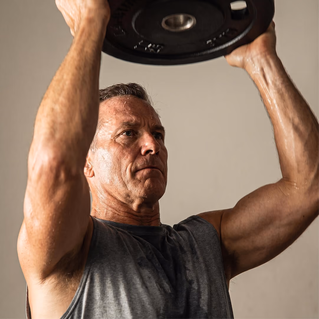 Muscular middle-aged man wearing a dark sleeveless shirt lifting a weight plate above his head with both hands, focused and sweating, against a plain background.