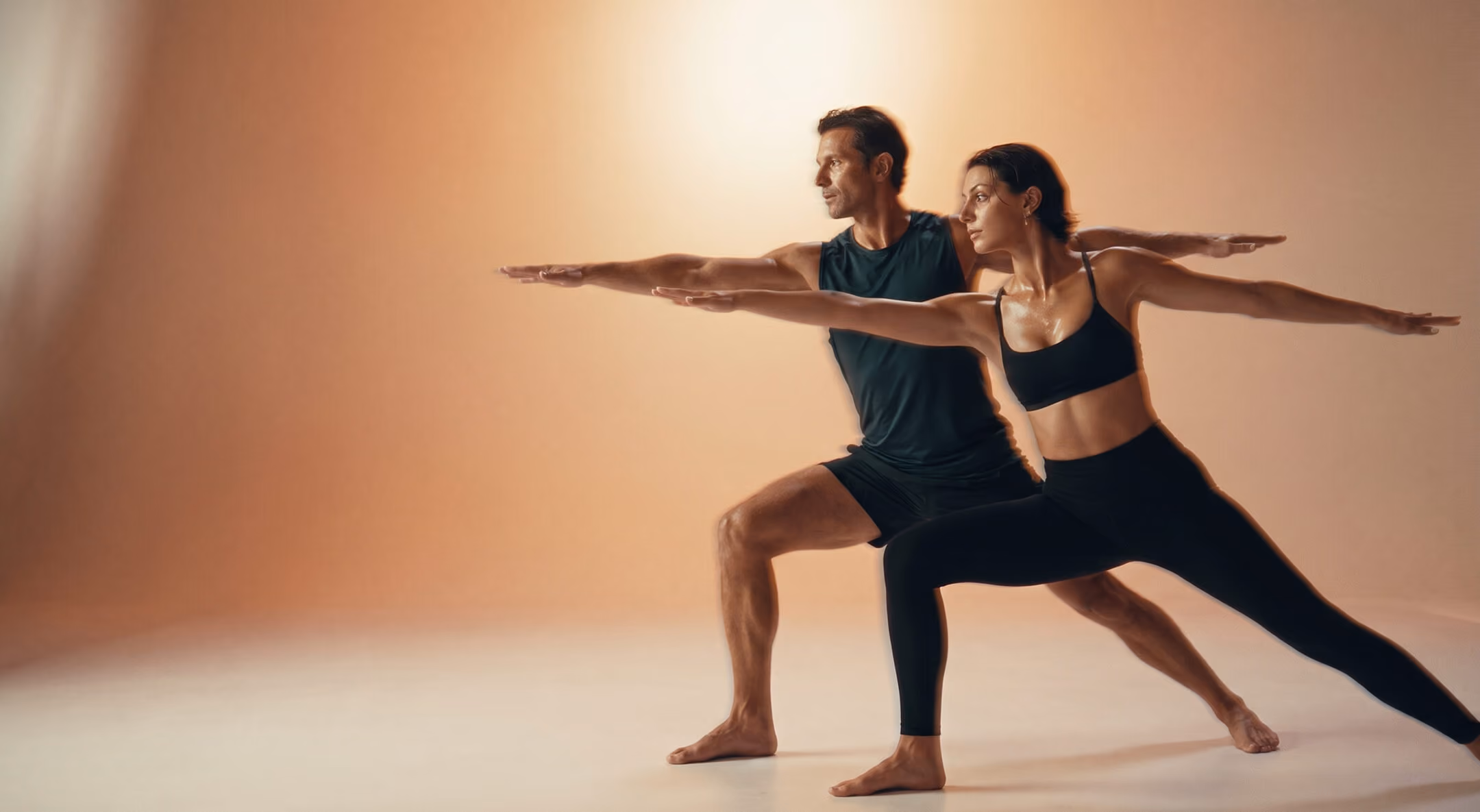 A man and a woman practicing yoga together in a studio, both in a Warrior II pose with arms extended and legs apart against a warm gradient background.