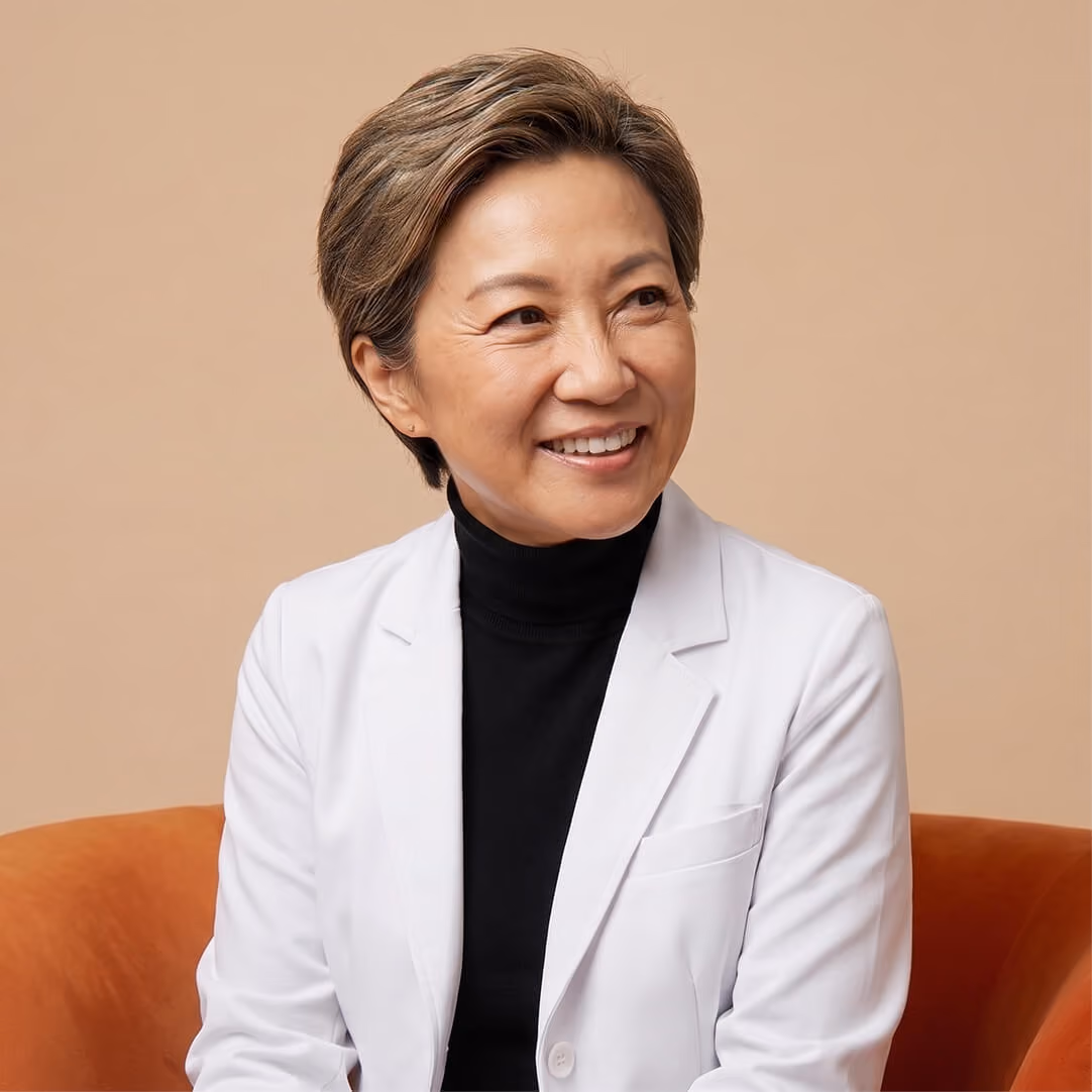 Smiling middle-aged woman with short hair wearing a white blazer over a black turtleneck seated on an orange chair against a beige background.