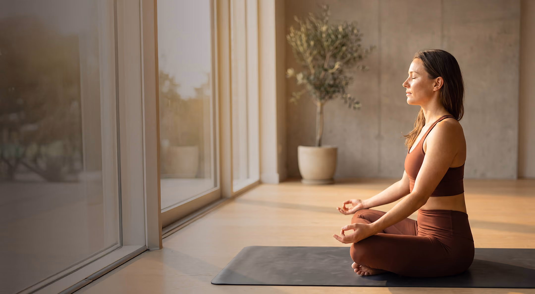 Woman in brown workout clothes sitting cross-legged on a yoga mat near large windows, meditating with eyes closed and hands resting on knees in a bright, calm room with a potted plant in the background.