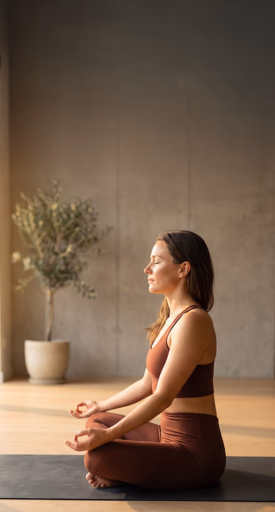 Woman in a brown sports outfit sitting cross-legged on a yoga mat with eyes closed, meditating indoors with a potted plant and plain wall in the background.