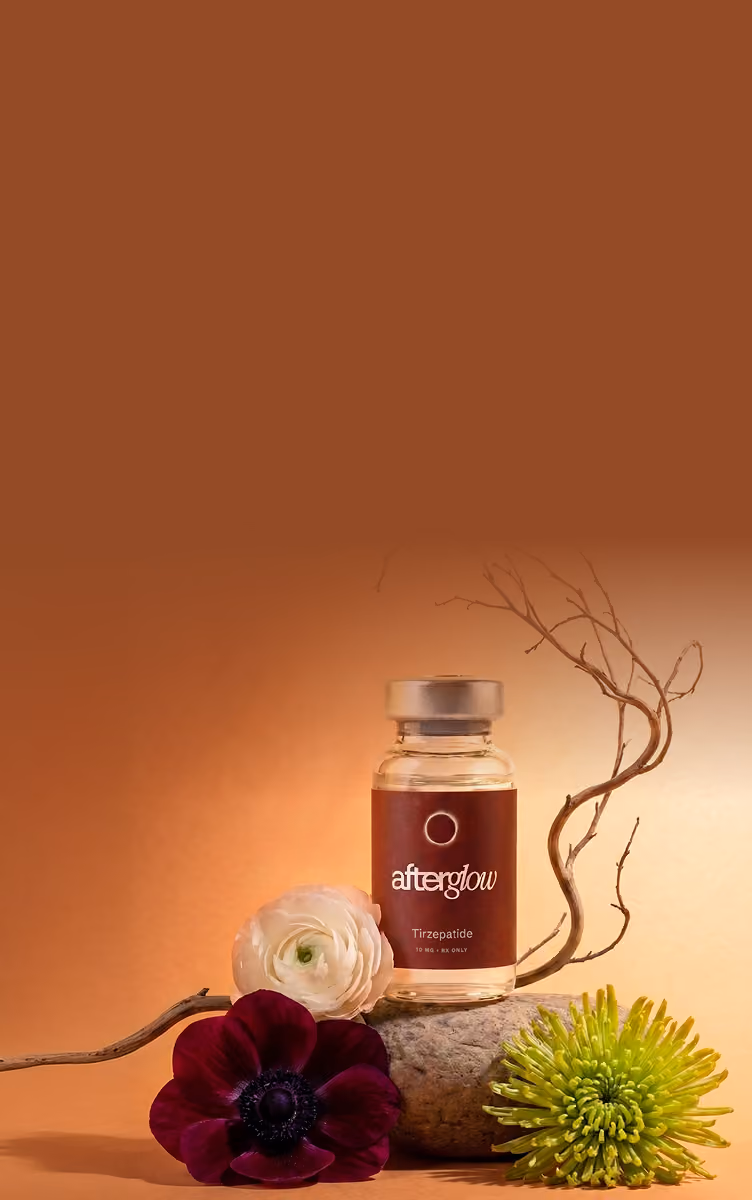 A small bottle labeled 'afterglow Tirzepatide' placed on a stone, surrounded by a white rose, a deep purple flower, a green chrysanthemum, and a thin curved branch against a warm brown to beige gradient background.