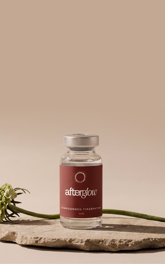 Clear vial labeled 'afterglow compounded tirazepatide 90 mg' with a maroon label, placed on a rough stone surface with a green flower stem in the background against a tan backdrop.