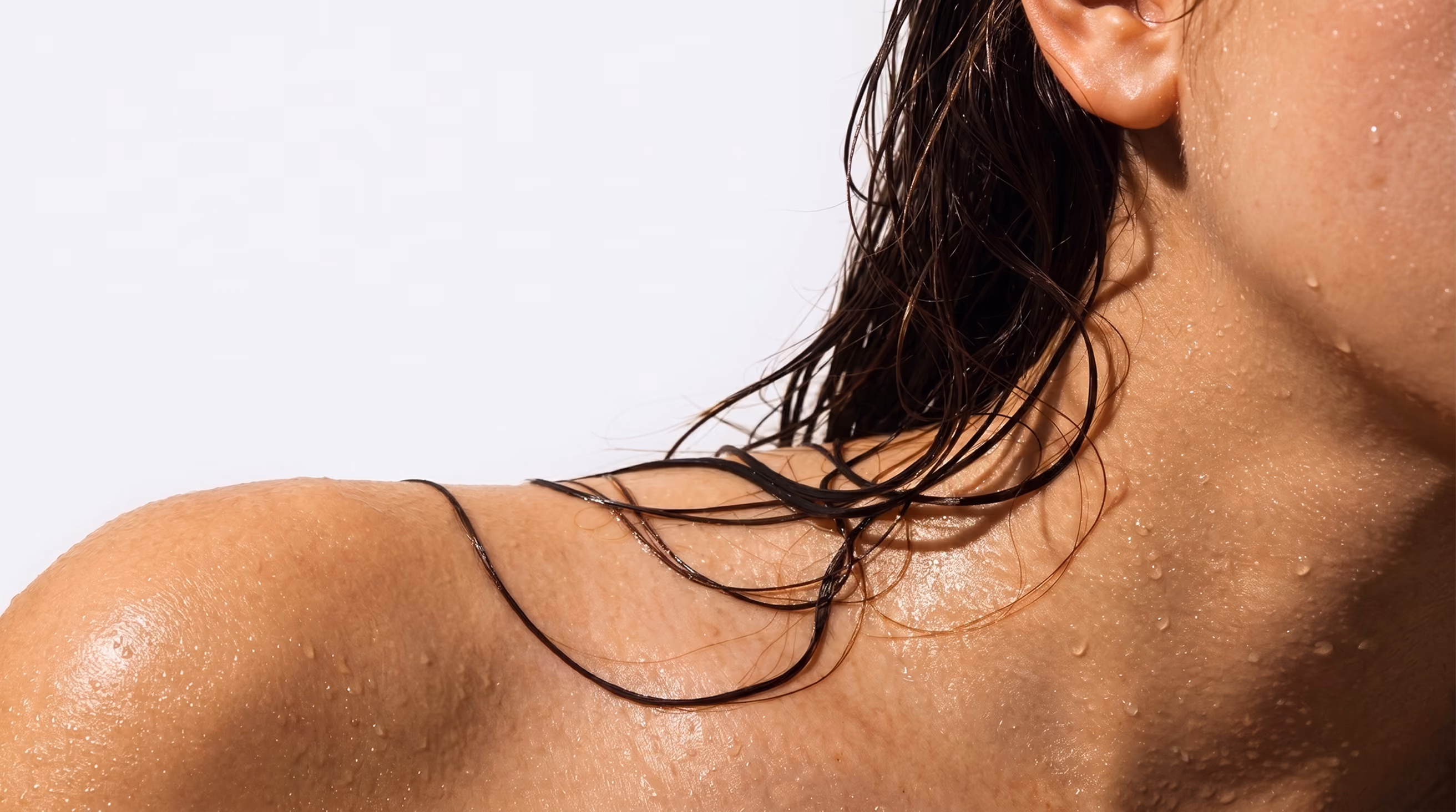 Close-up of a woman's wet neck and shoulder with strands of dark wet hair resting on her skin against a light background.