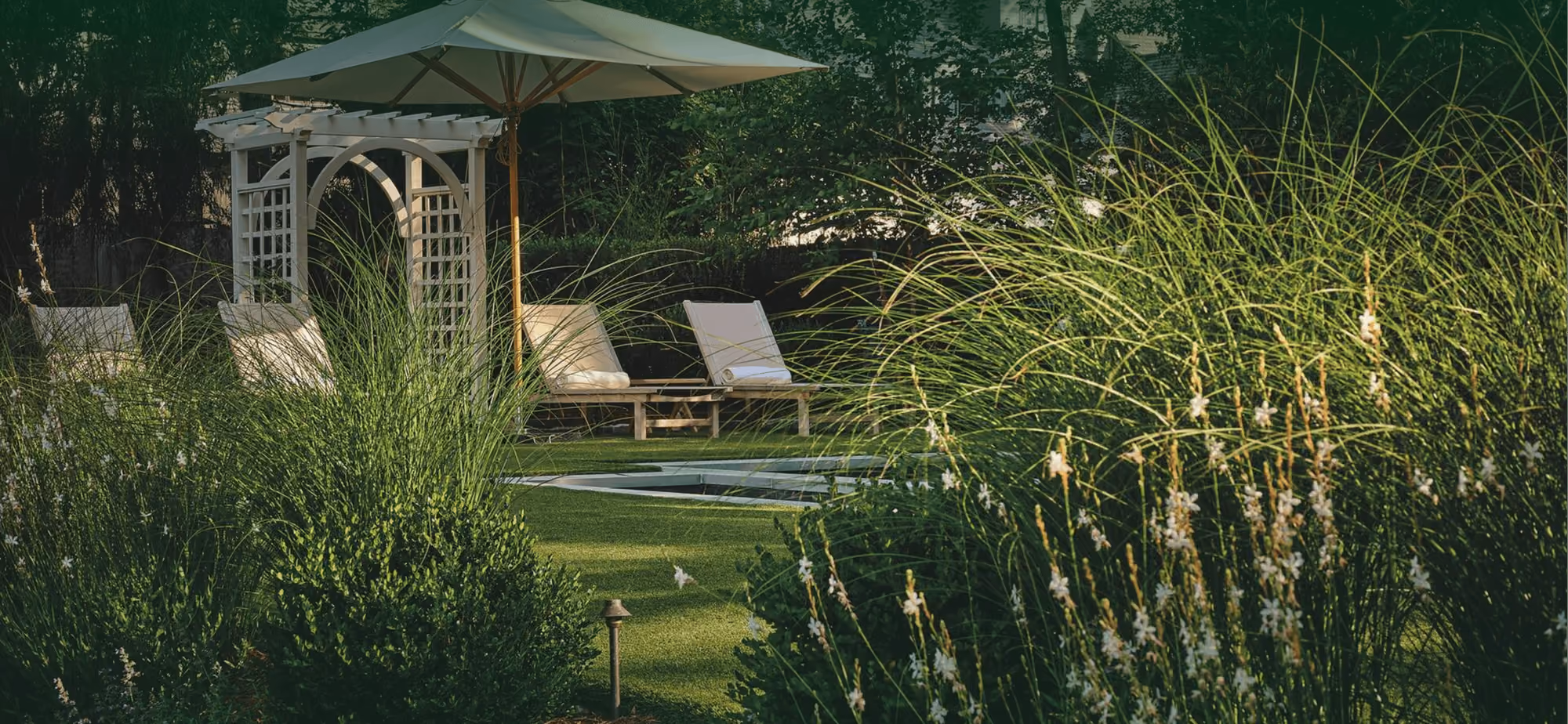 Outdoor garden scene with two lounge chairs under a large patio umbrella near a round hot tub, surrounded by lush green bushes and tall grasses.