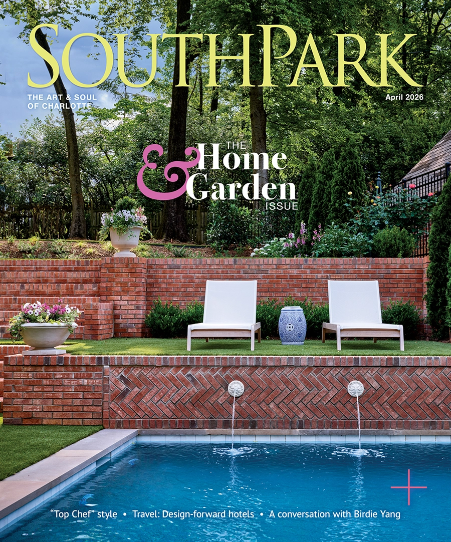 Outdoor pool area with clear blue water in front of a red brick wall featuring two water spouts, two white lounge chairs, a blue decorative side table, and potted flowers, set against a backdrop of green trees under a blue sky.