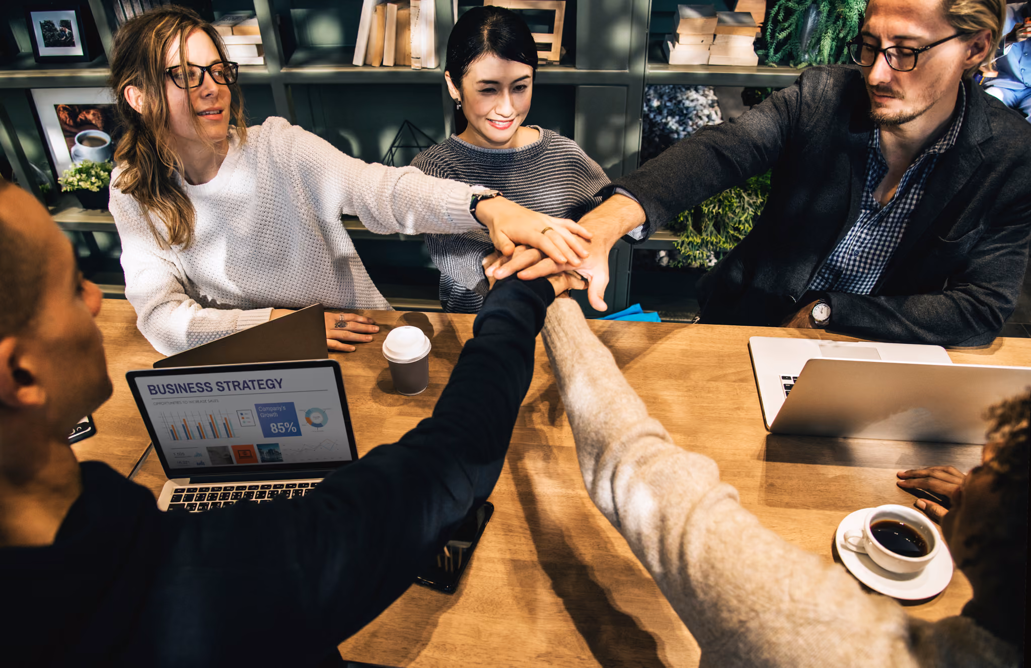 Five diverse colleagues sitting around a wooden table join hands in a team gesture, with laptops, a coffee cup, and a disposable cup on the table.