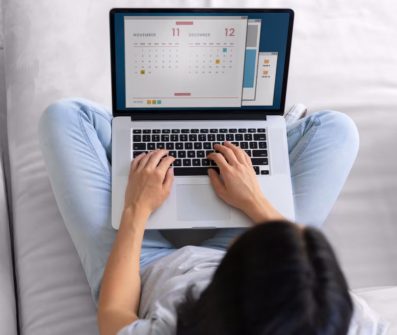 Person sitting cross-legged on a couch using a laptop showing calendar months November and December on the screen.