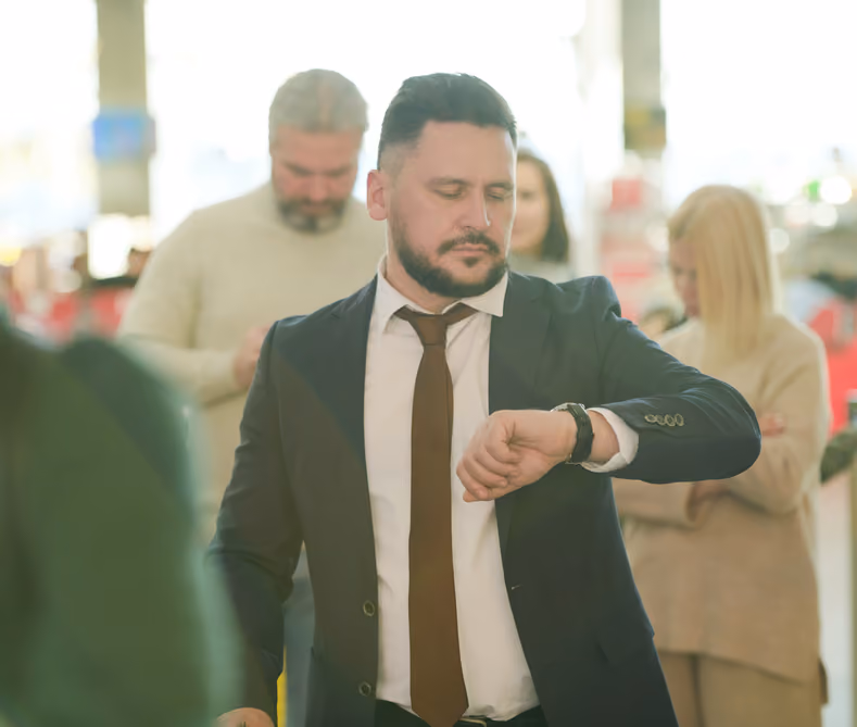 Man in a suit with a brown tie looking at his wristwatch in a busy indoor setting with blurred people in the background.