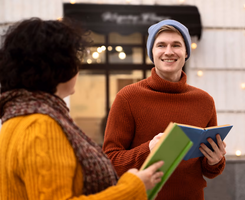 Two people holding open books and smiling, one wearing a red sweater and blue beanie, the other in a yellow sweater with a scarf, standing outdoors in front of a building.