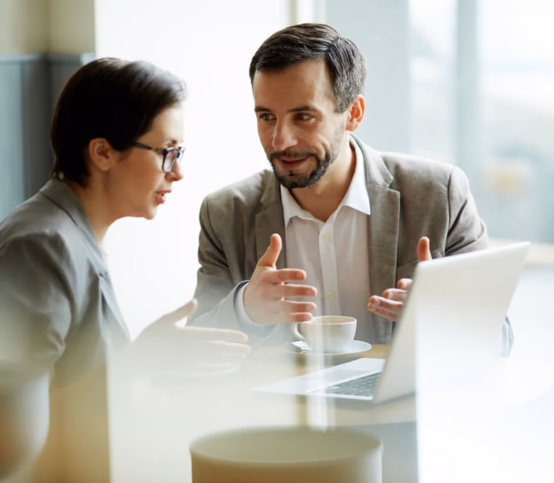 Two business professionals, a man and a woman, engaged in a discussion while sitting at a table with a laptop and a cup of coffee.