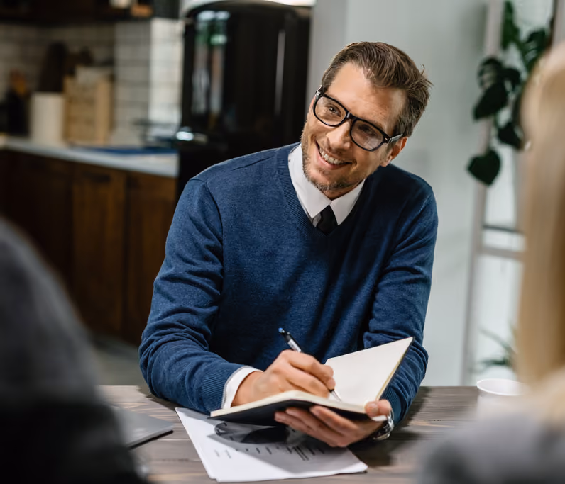 Smiling man wearing glasses, a blue sweater, and a tie, sitting at a table and writing in a notebook during a conversation.
