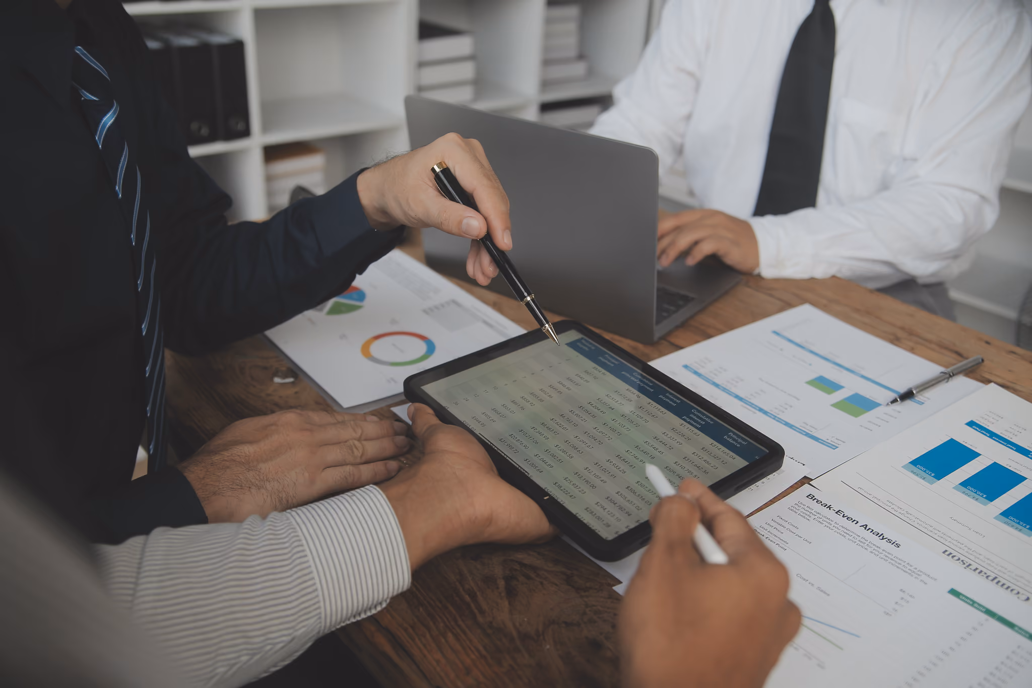 Three businesspeople at a wooden table analyzing financial data and charts displayed on a tablet, laptop, and printed papers.