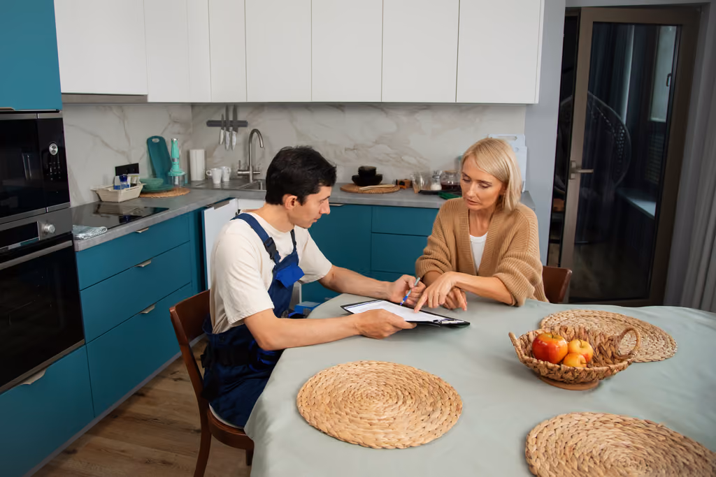 A man in work overalls sitting at a kitchen table discussing documents with a woman wearing a beige sweater, pointing at the papers.