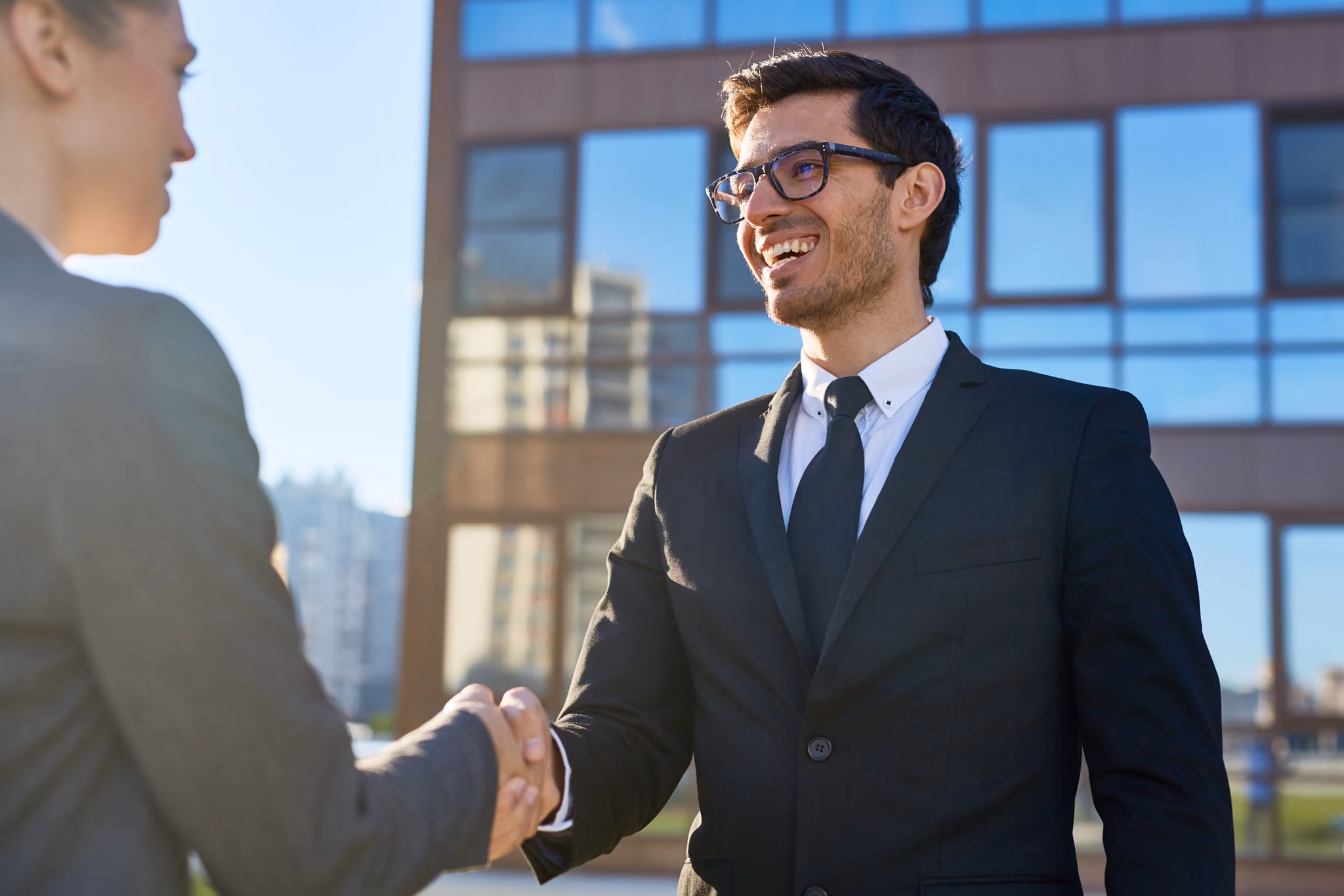Businessman in a black suit and glasses smiling and shaking hands with a woman in a black blazer outside an office building.