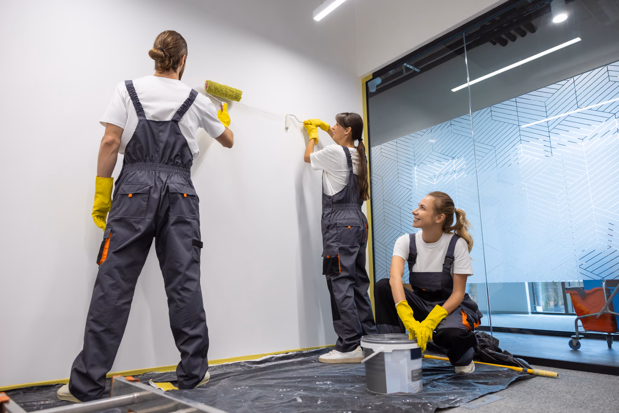 Three workers in white shirts and gray overalls wearing yellow gloves painting a white wall indoors, one using a paint roller, another holding a smaller roller, and the third sitting next to a paint bucket and smiling.