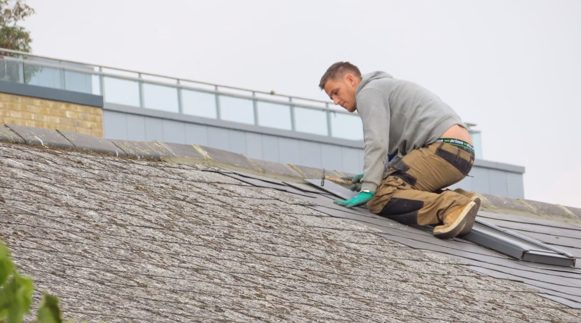 A man wearing a gray hoodie, tan work pants, green gloves, and brown boots is kneeling on a shingled roof, installing or repairing black roof tiles while holding a hammer.