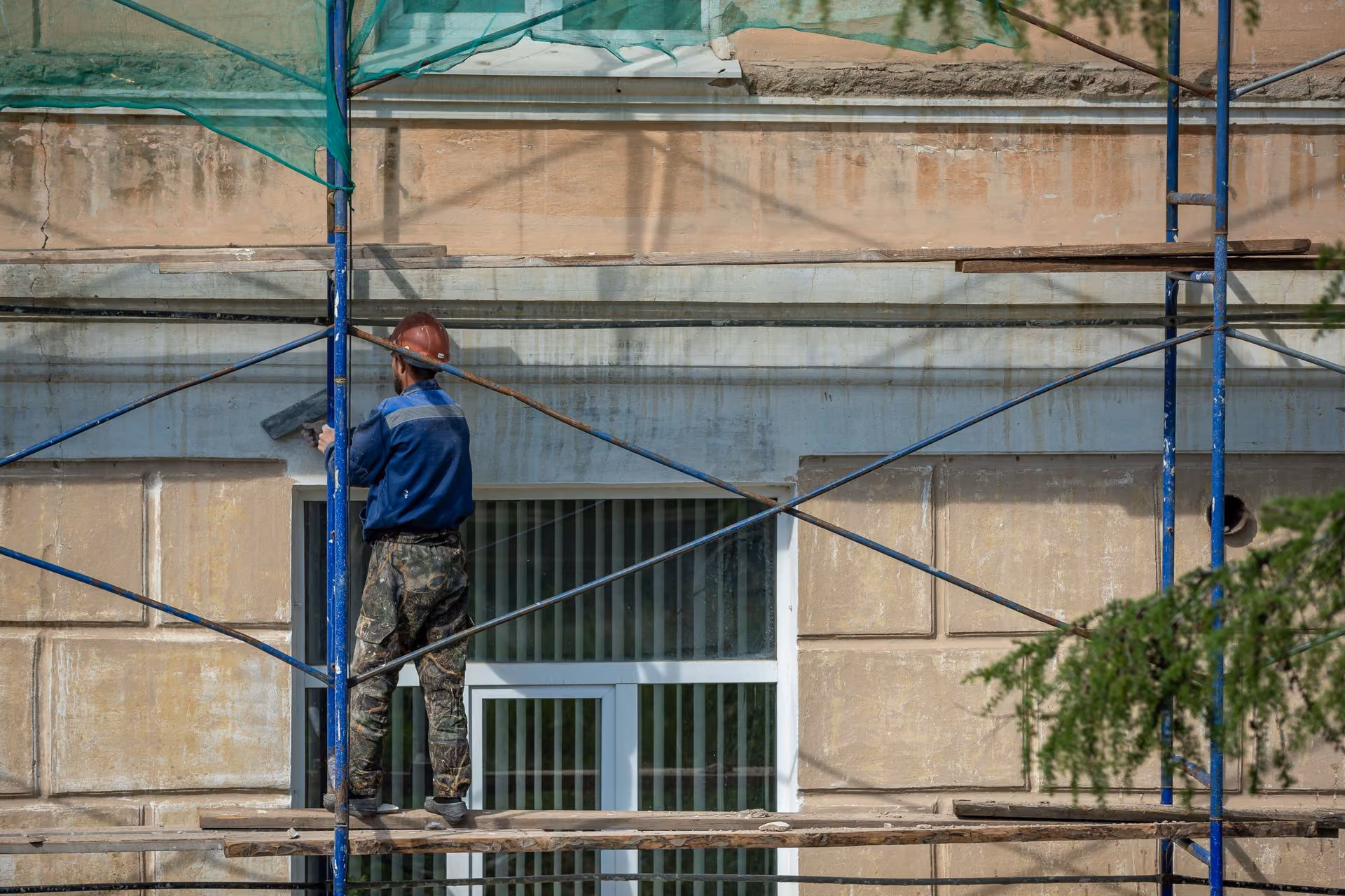 Construction worker in a blue shirt, camouflage pants, and red helmet applying plaster or cement to a building wall through scaffolding.