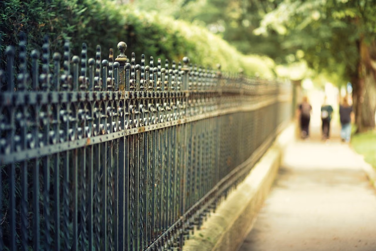 Long black wrought iron fence running alongside a sidewalk with green bushes behind it and blurred figures of three people walking in the distance under tree shade.