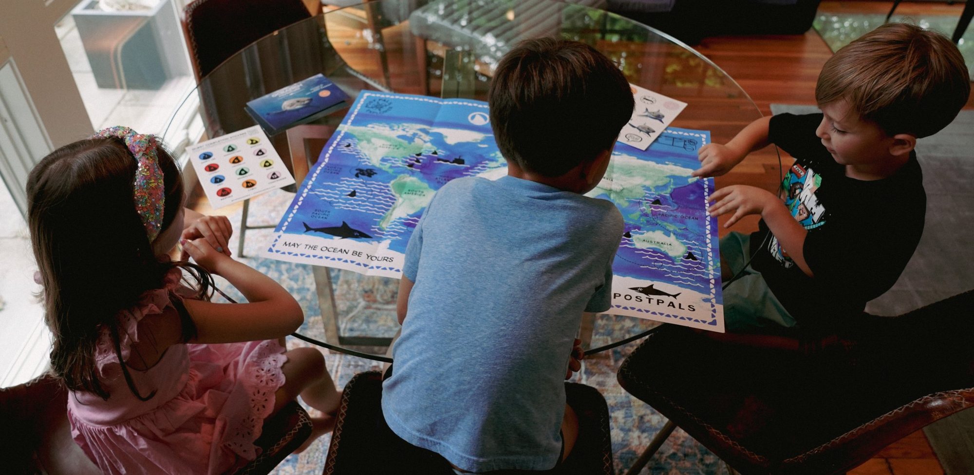 Three kids lean over a POSTPALS ocean map spread across a glass table at home