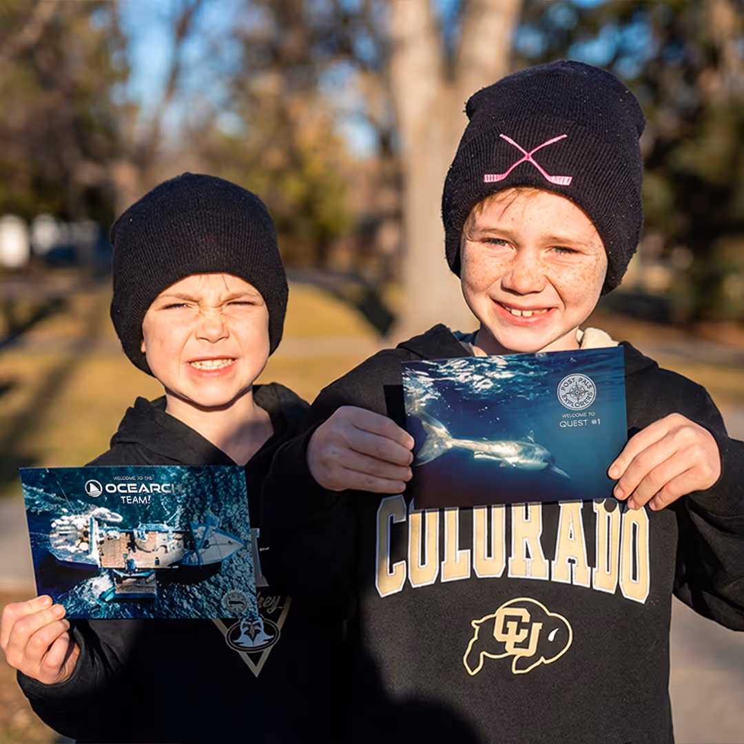 Two boys wearing black beanies and hoodies holding POSTPALS postcards.