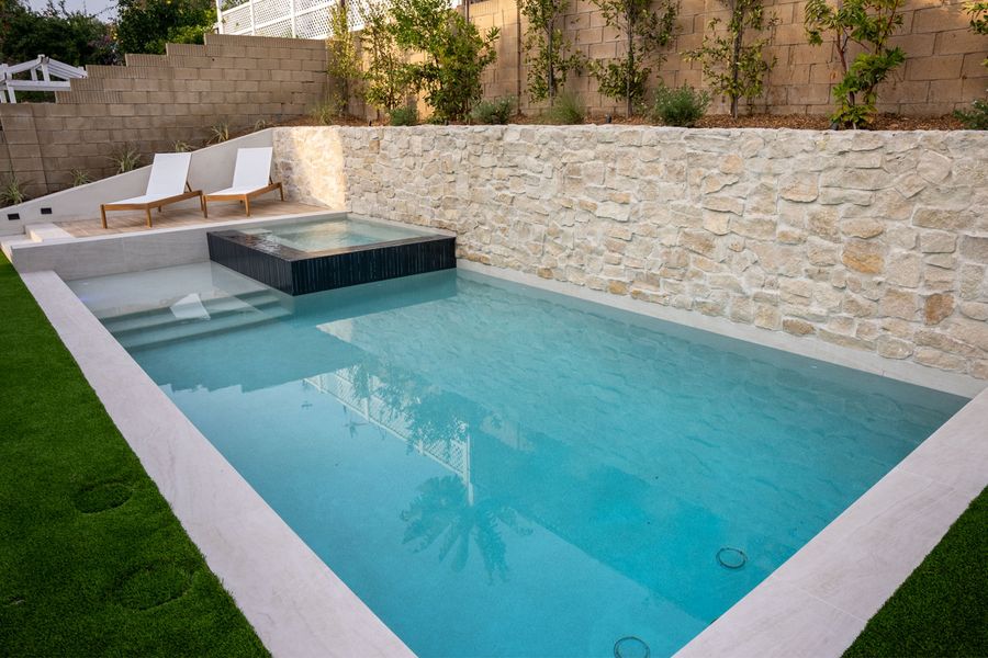 Pale stone coping against turquoise water, raised spa at the far end, stone wall behind.
