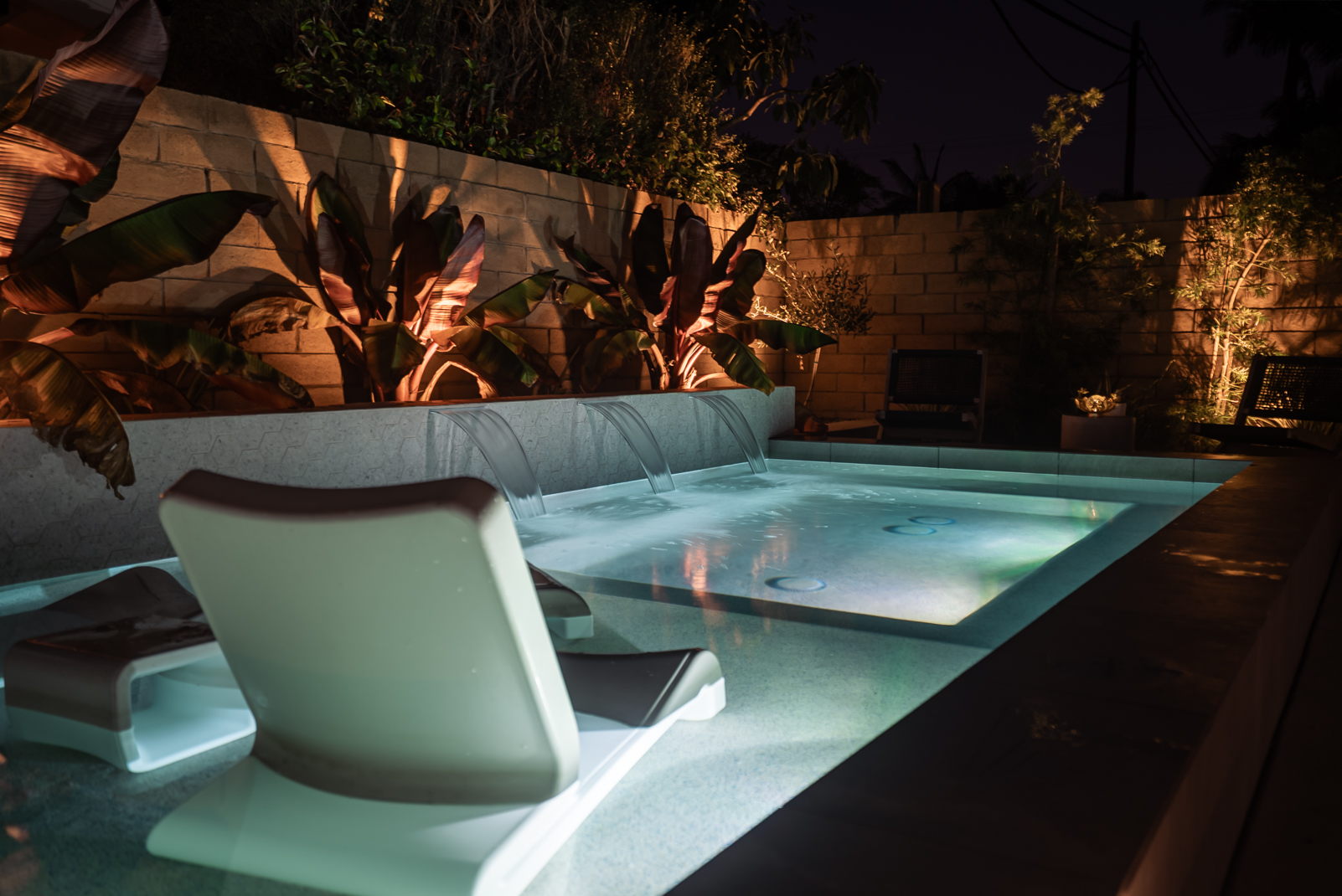 A compact rectangular pool with a Baja shelf, pale coping, turf, a lounge chair, and a stacked-stone garden wall.