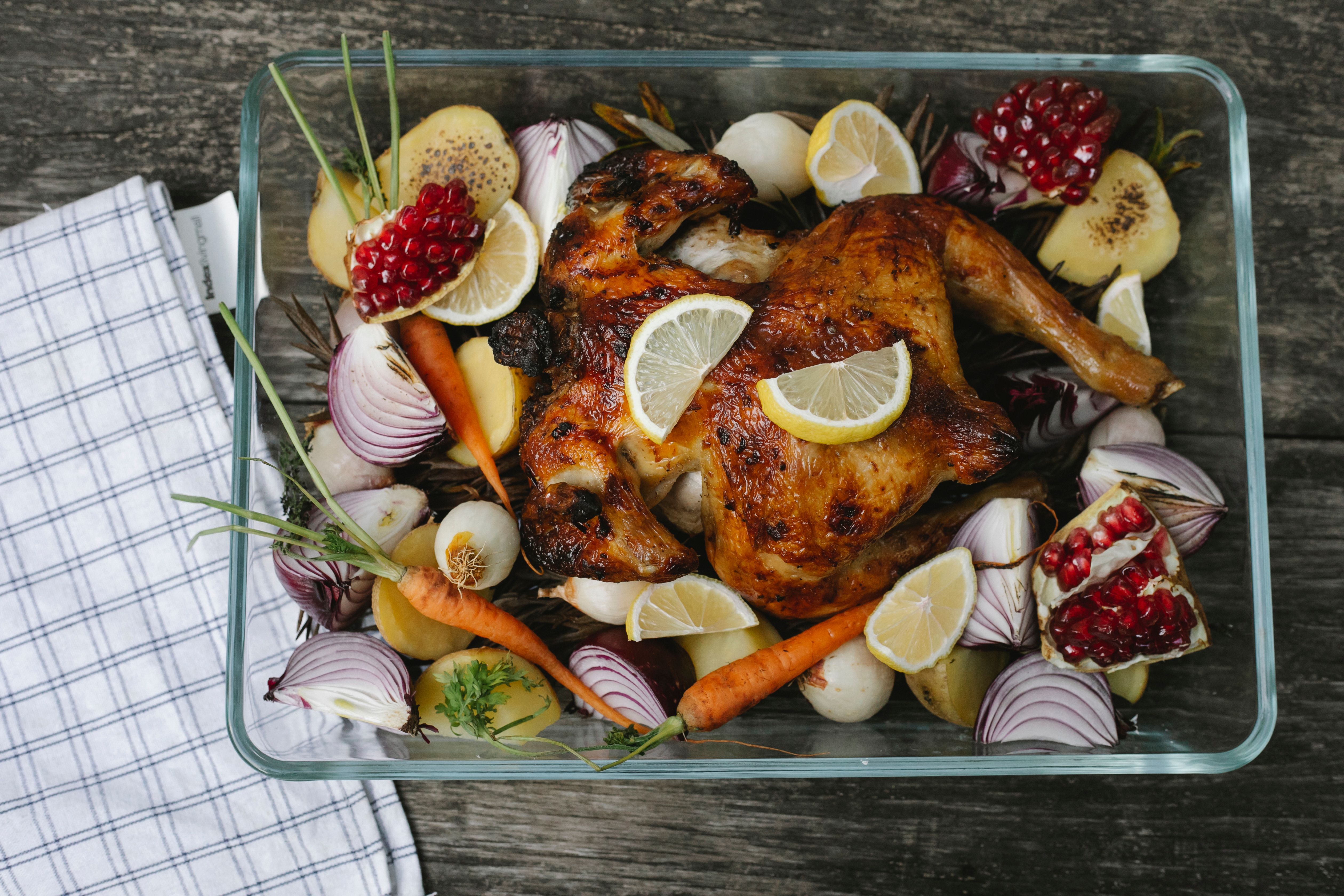 A roast bird in a glass tray surrounded by carrots, red onion, potatoes, lemon and pomegranate.