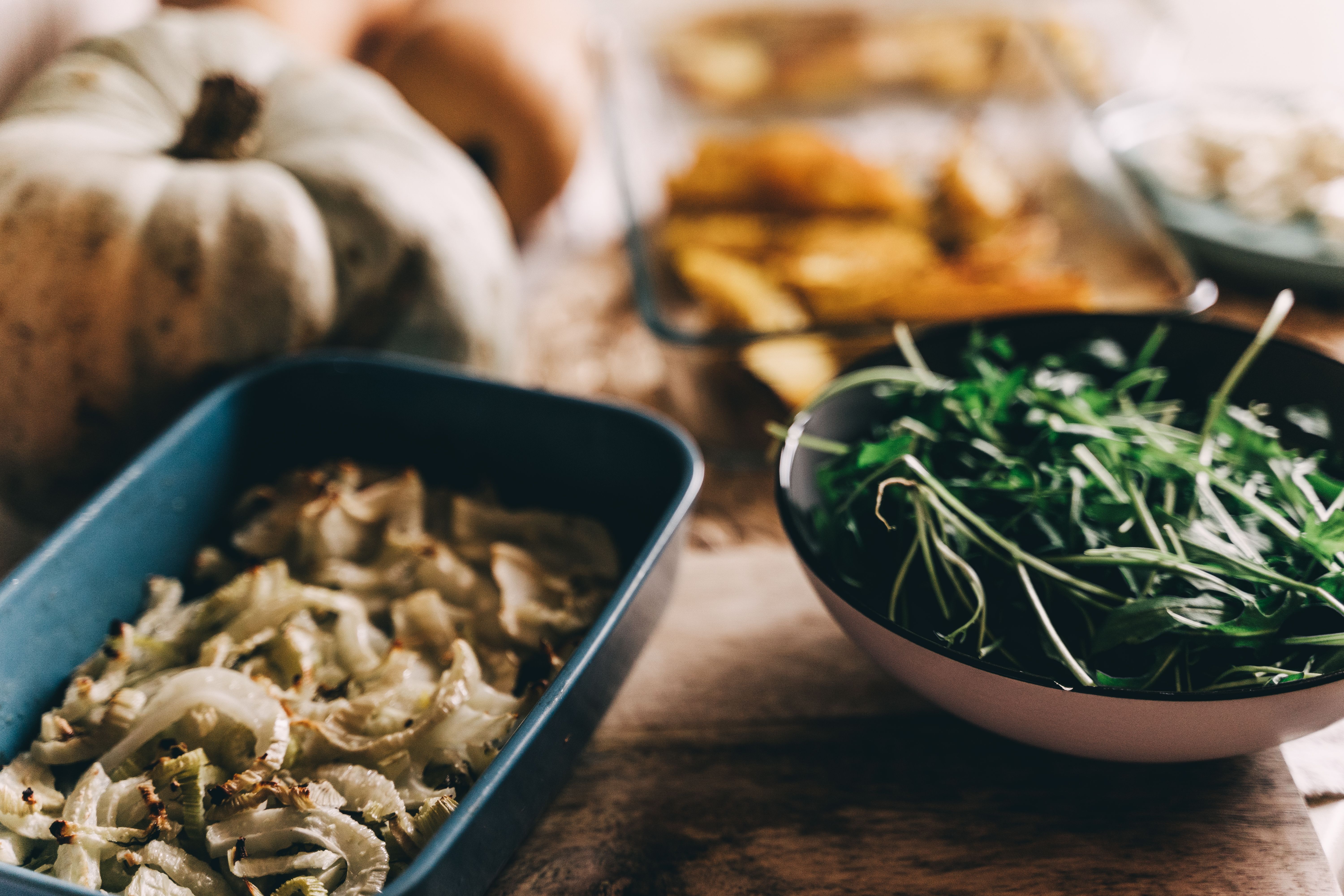 Holiday side dishes on a wooden table: roasted fennel, potatoes and a bowl of fresh greens beside a white pumpkin.