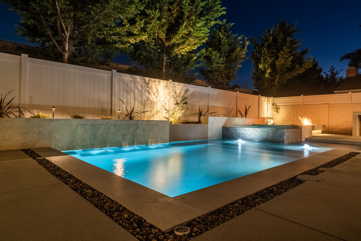 The whole pool at blue hour, a dark coping band around the water and trees uplit along the fence.