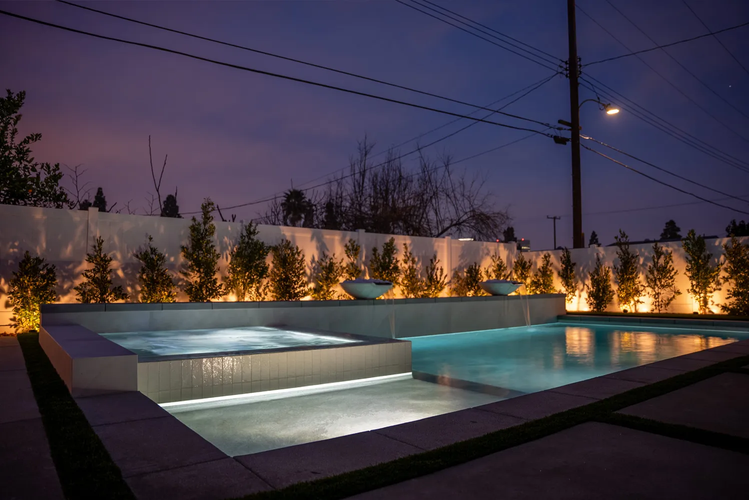 A wide dusk view of the pool and spa, the row of trees along the wall lit from below.