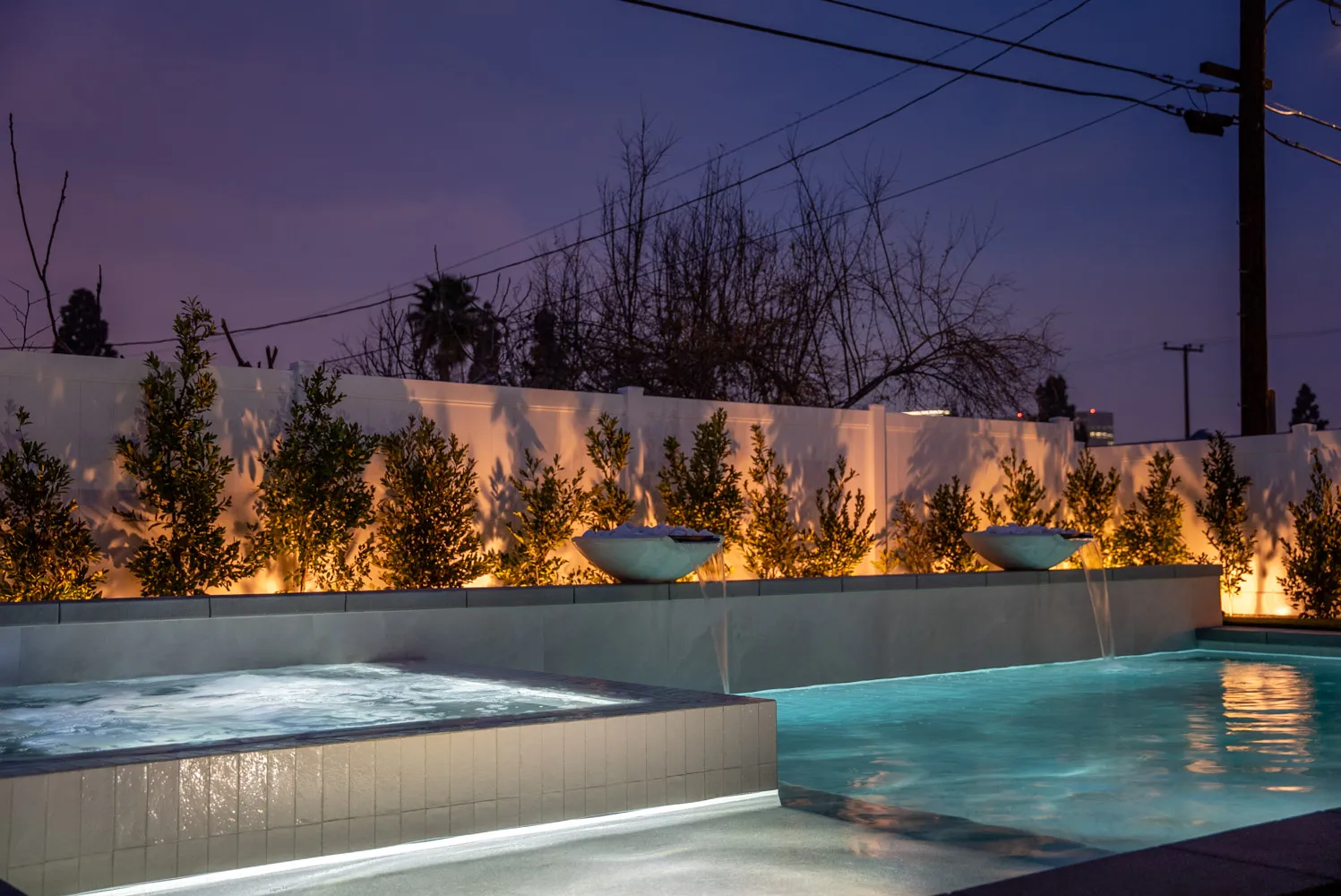 The spa steaming at dusk under a violet sky, with the planted wall uplit behind and water bowls spilling into the pool.