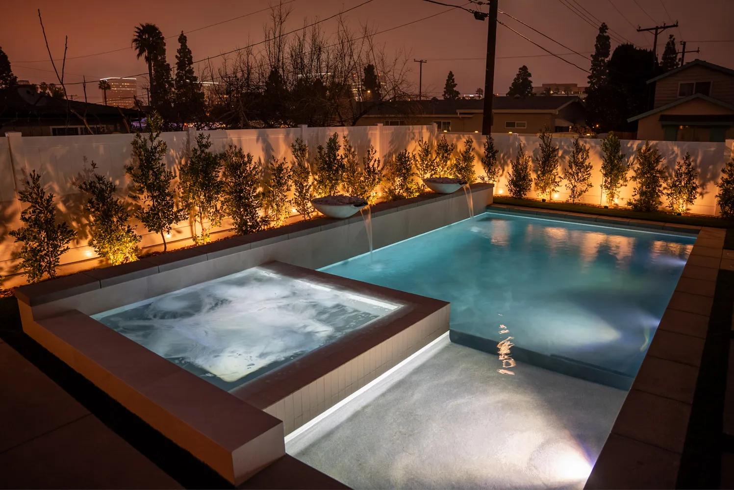 The spa and pool from above at sunset, the uplit hedge running behind and an orange sky over the rooflines.
