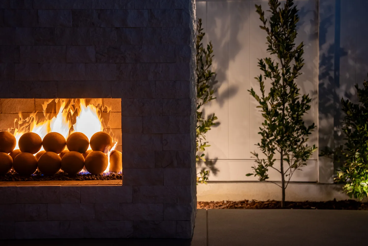 The fireplace burning at night, its light falling across a young tree and the pale boundary wall.