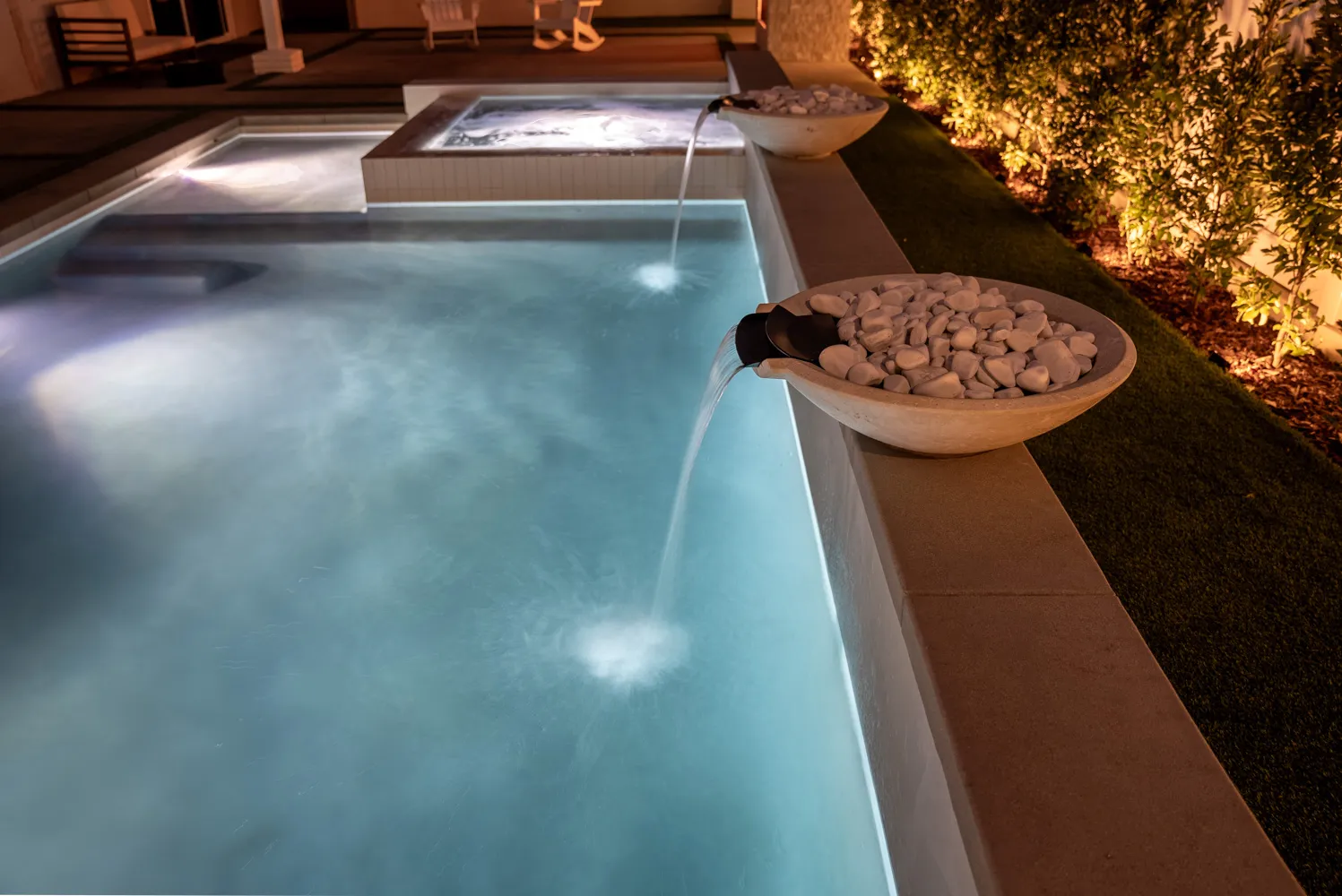 A bowl pouring into the lit pool after dark, the spa glowing at the far end.