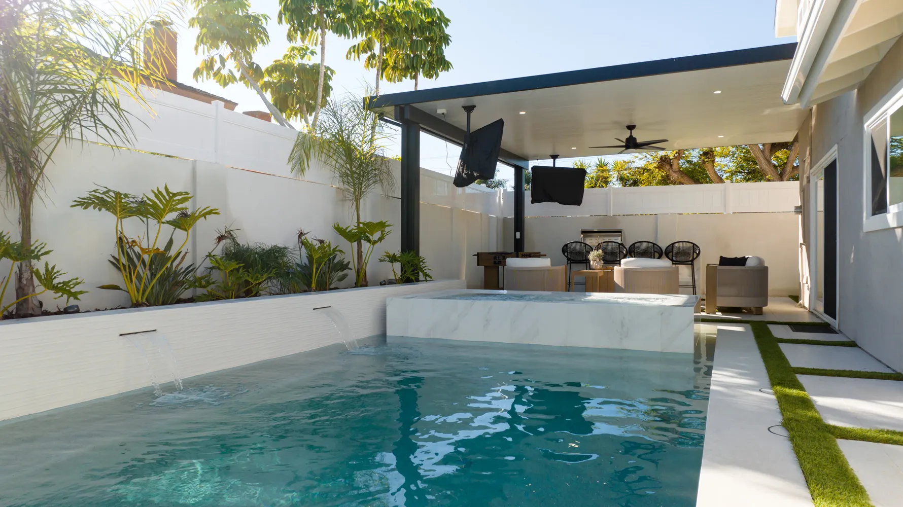 The pool in full sun with a covered lounge along the far side, palms and tropical planting rising over a white wall.