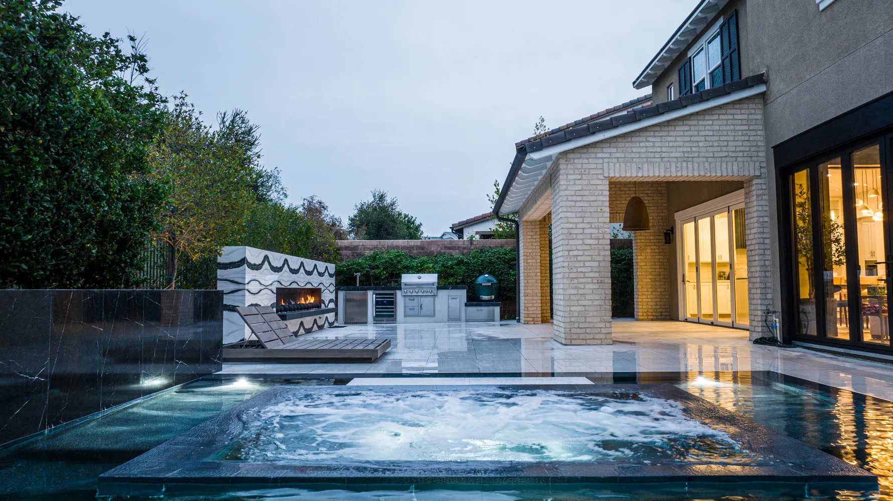 The pool at dusk with the house lit behind it, a raised spa in the foreground and a long fireplace burning across the far deck.