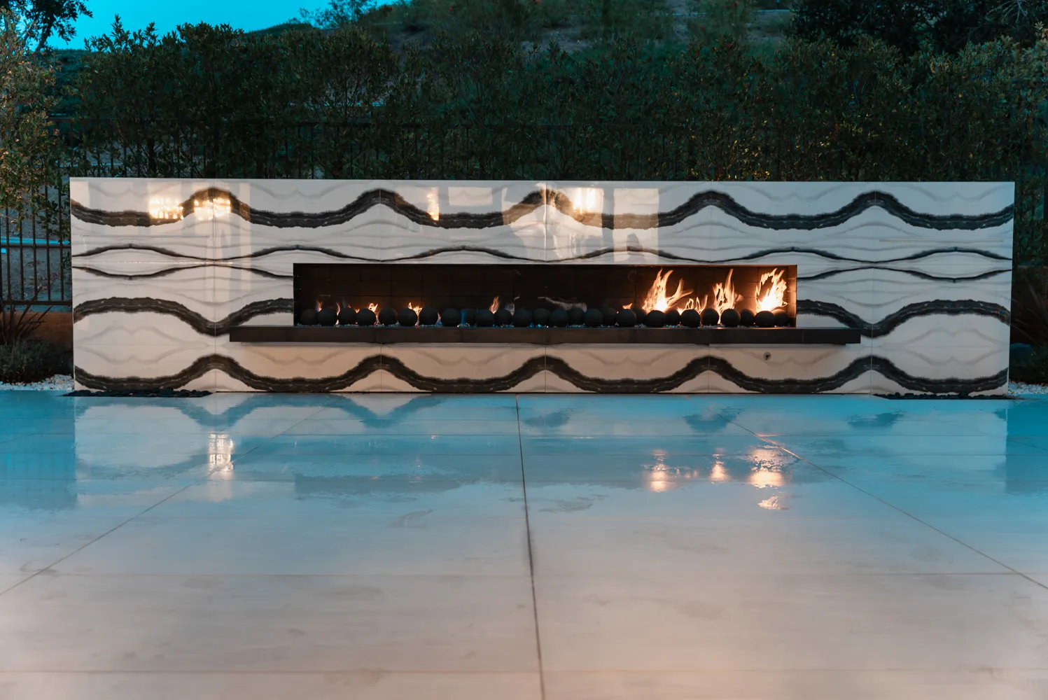 A long linear fireplace burning in a wall of figured marble, its light reflected across the wet deck.