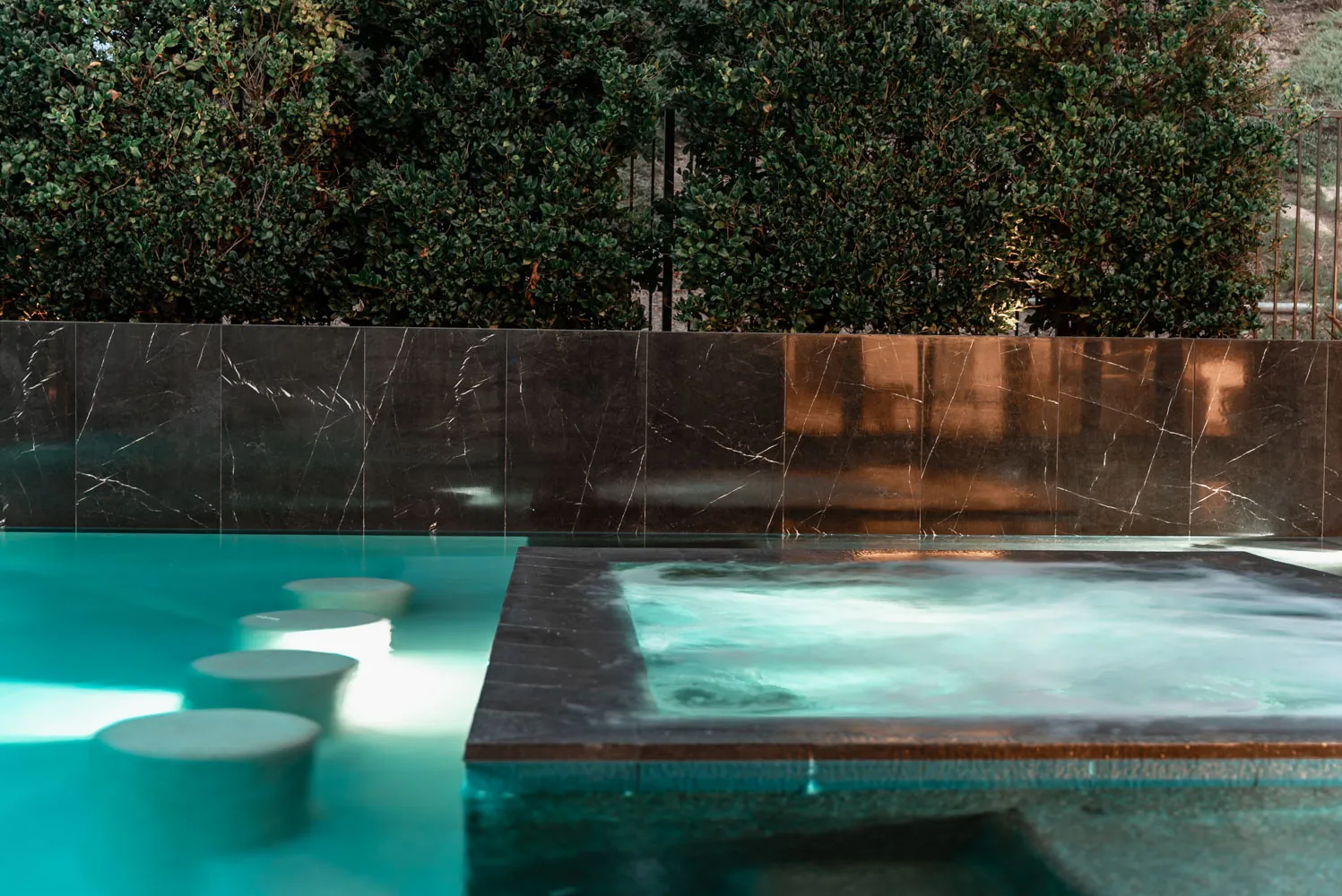A raised spa clad in dark marble, lit from within, with pale stools standing in the shallow water beside it.
