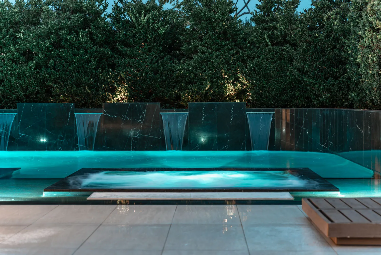 Sheet waterfalls dropping into the lit pool at dusk, a timber deck running along the near edge.