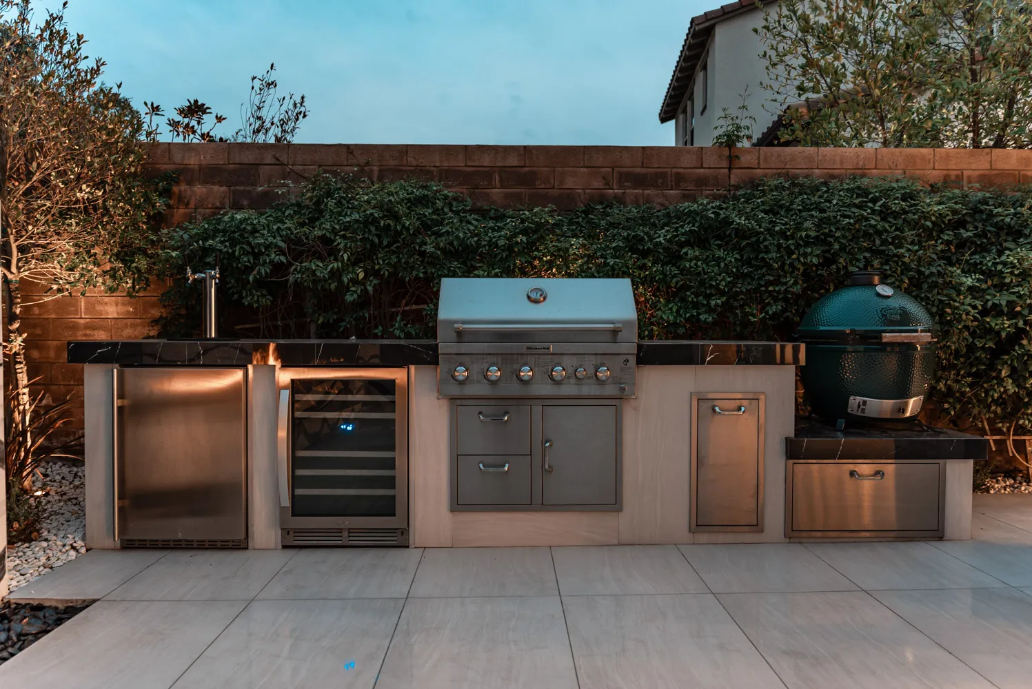 The outdoor kitchen at dusk, with a grill, wine refrigerator and ceramic smoker set into a dark stone counter against a hedge.