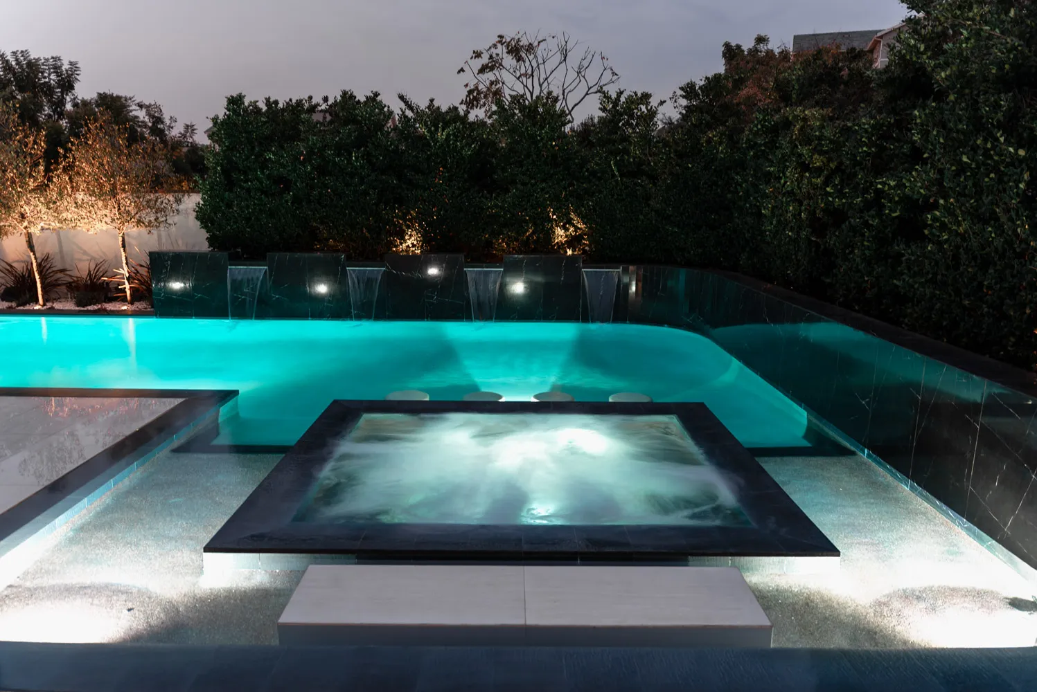 A raised spa in a dark tile surround, sheet falls behind it and a hedge closing the far boundary.