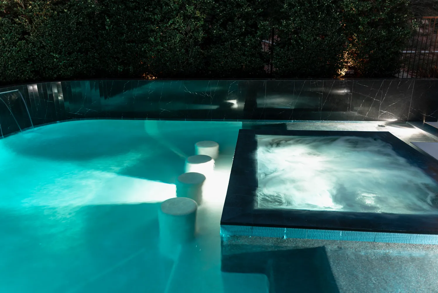 The spa spilling into the pool after dark, pale stools set in the shallow water.