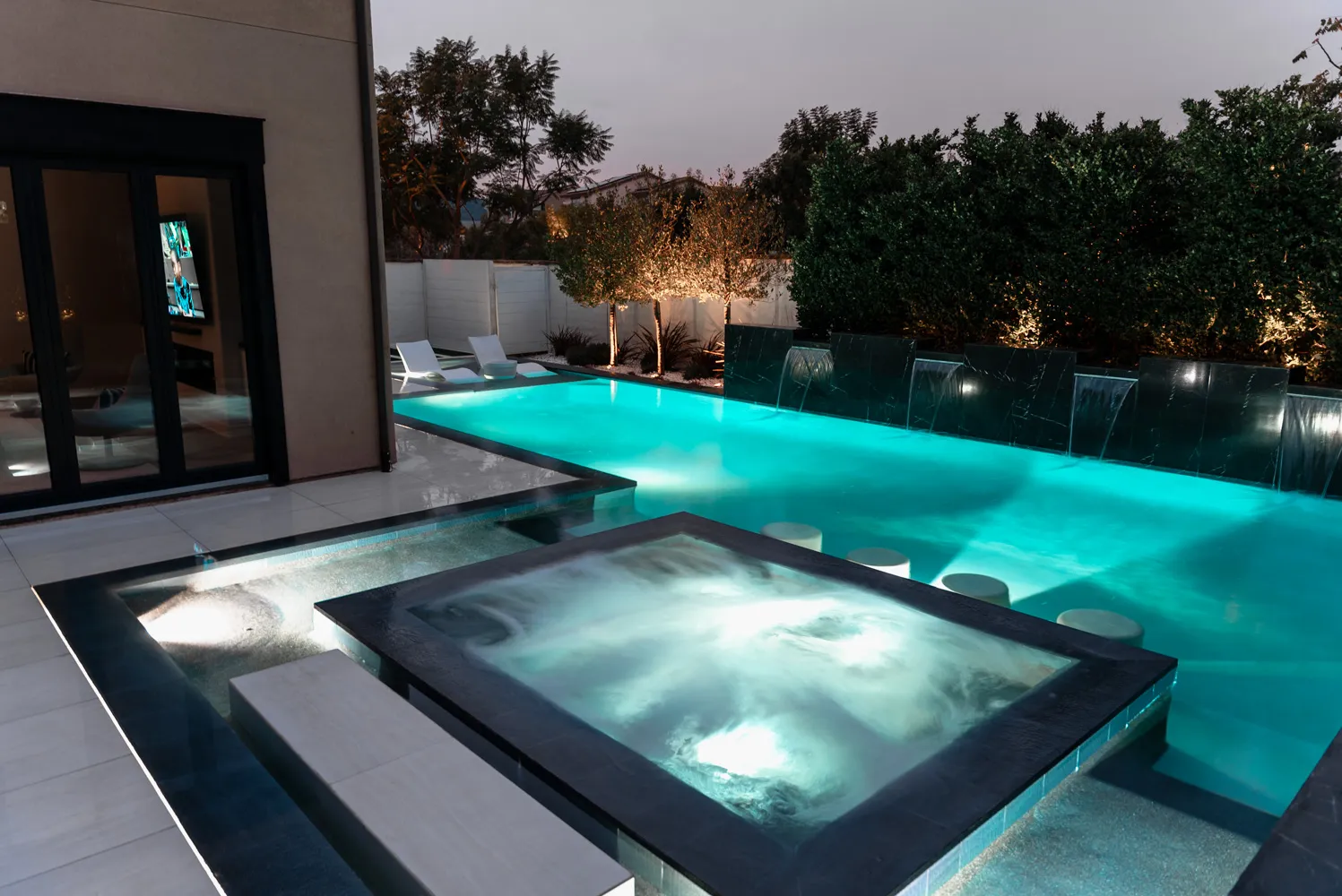 The pool and spa after dark seen from the terrace, the lit interior of the house showing through the glass.