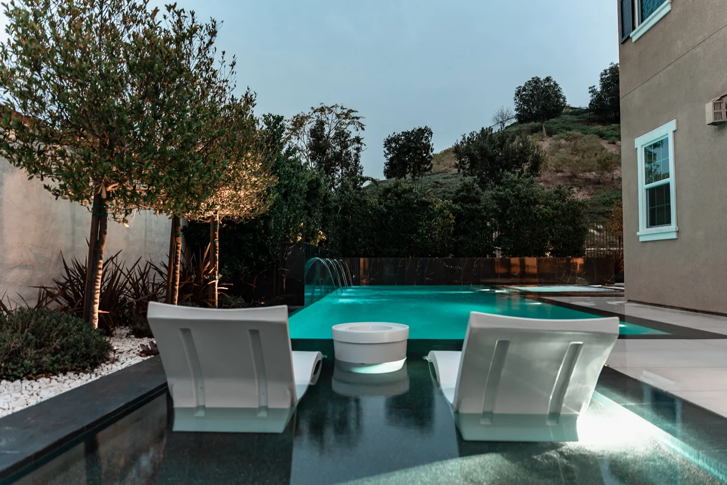 Two loungers standing in the shallow end at dusk, the house rising behind the deck.