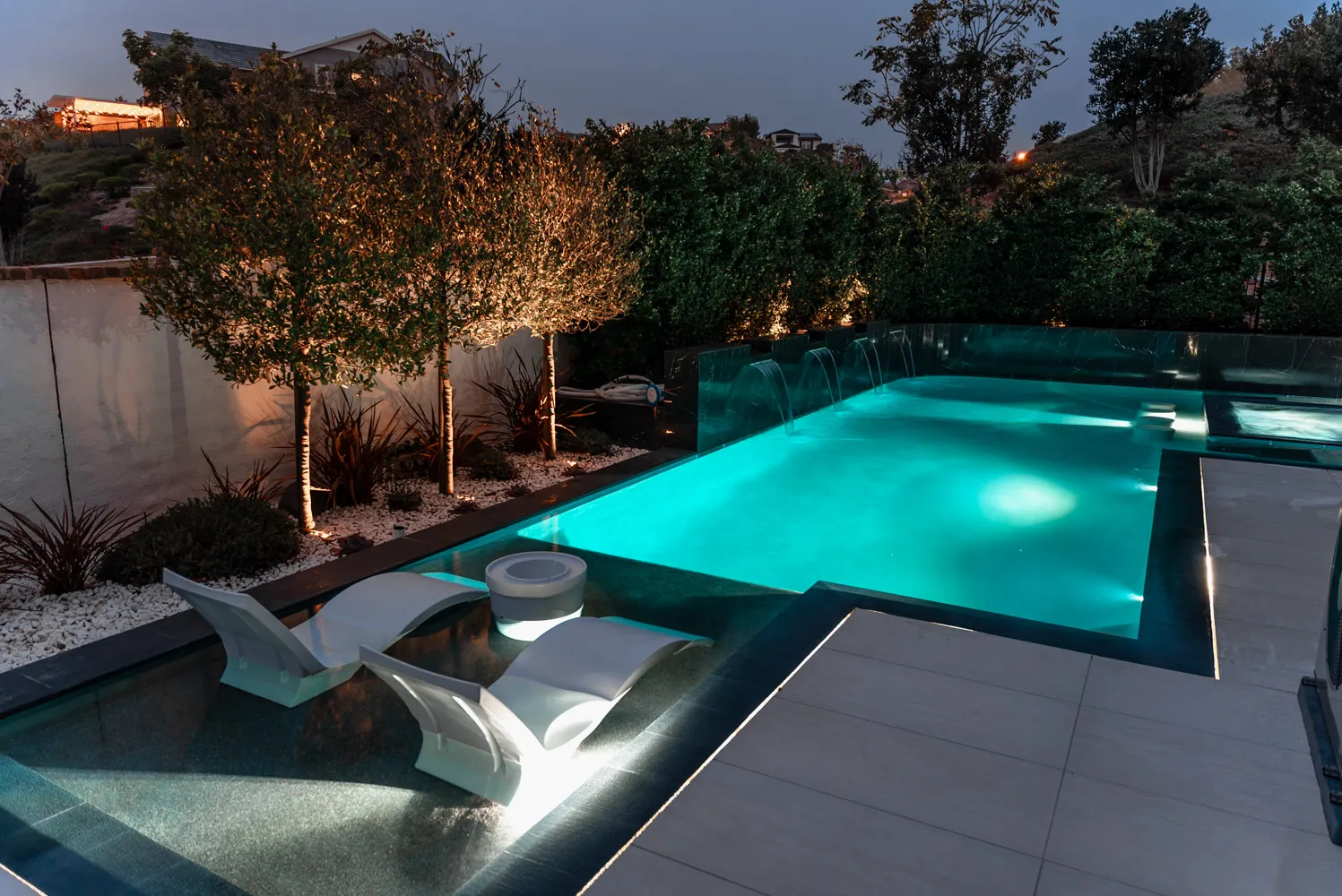 The lit pool at dusk with loungers standing in the shallow end and young trees uplit along the deck.
