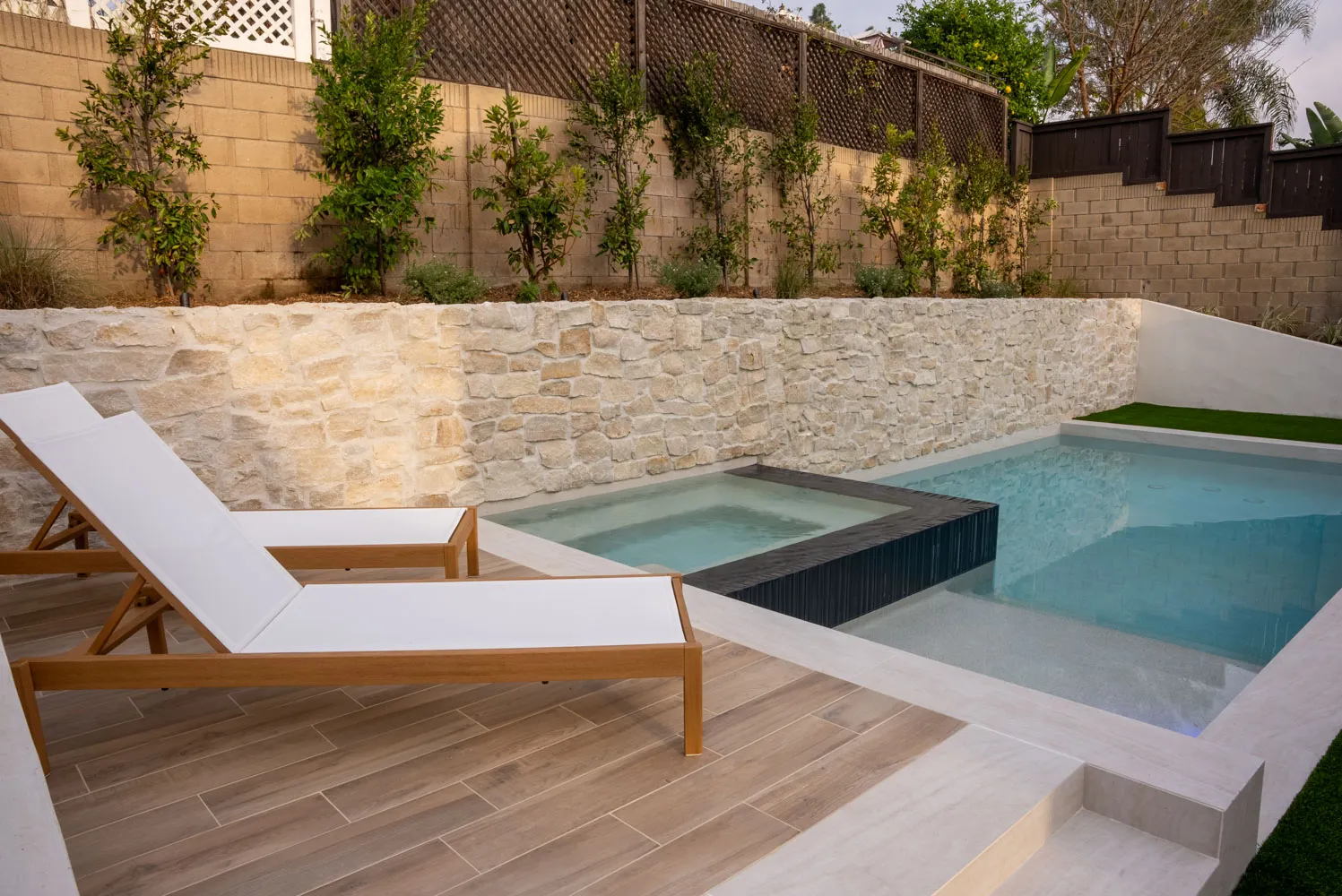 Two teak loungers on a timber deck beside a dark-tiled spa, with a stacked-stone retaining wall rising behind.