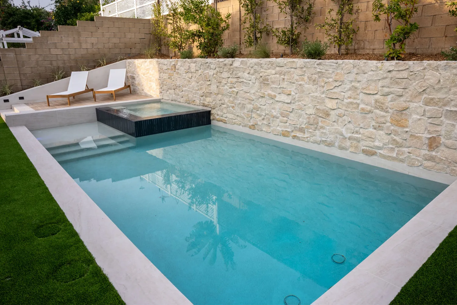 The full rectangle of the pool from its corner, turf on one flank and a stacked-stone wall closing the far side.
