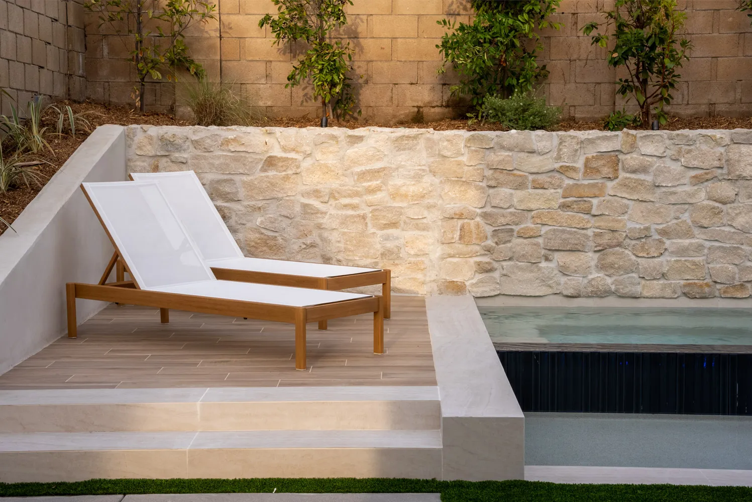 A single lounger on pale paving beside the spa, the stone wall running the length of the frame.